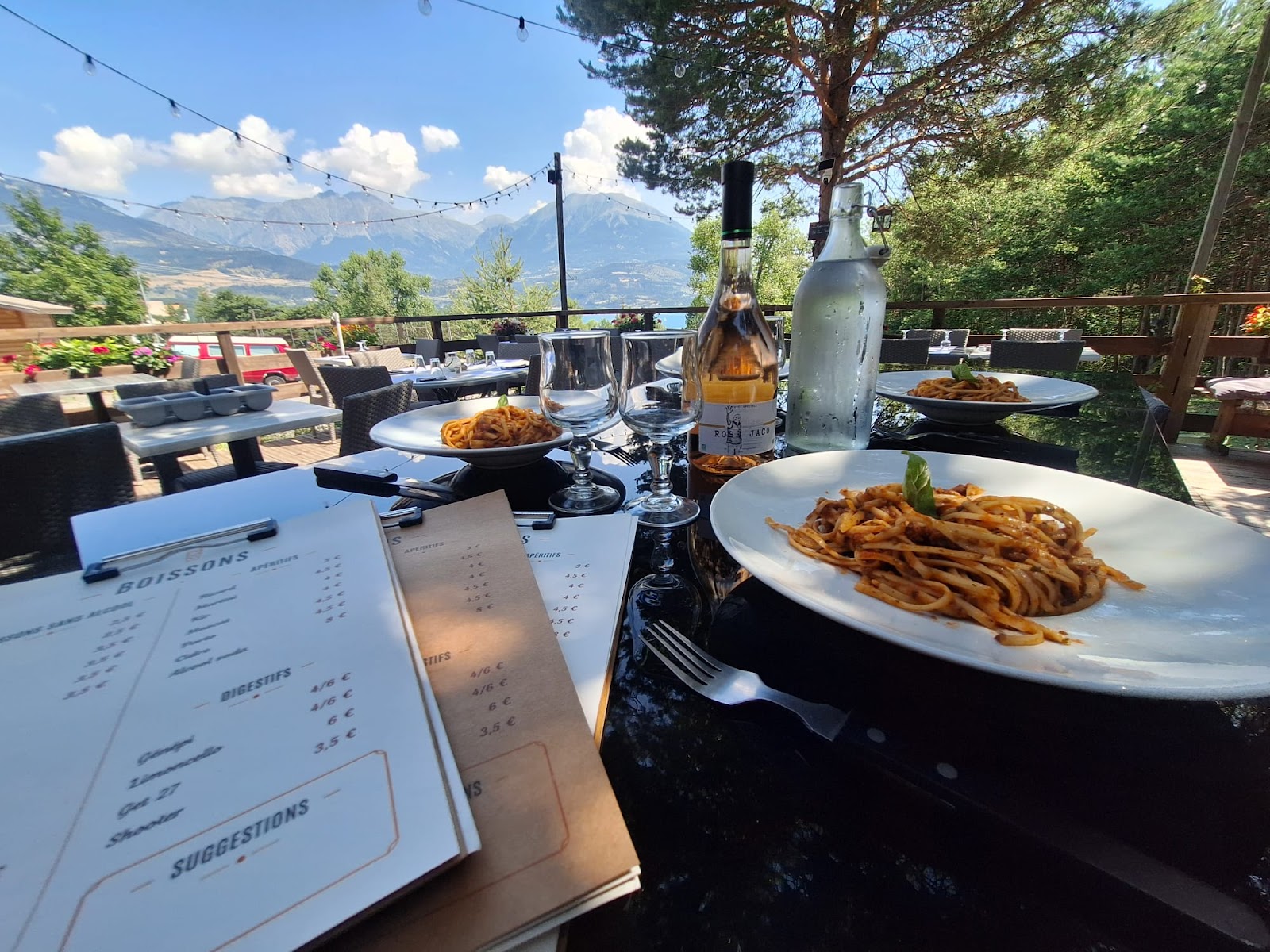 Plats de pâtes, bouteille de rosé et ardoise sur la table en terrasse, vue sur les montagnes